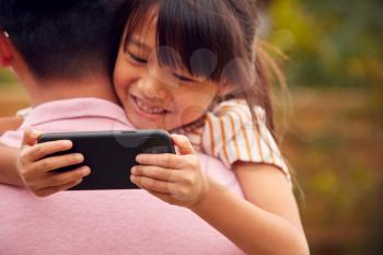 Asian Father Cuddling Daughter In Garden As Girl Looks Over His Shoulder At Mobile Phone