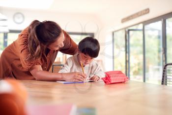 Asian Mother Helping Home Schooling Son Working At Table In Kitchen Writing In Book
