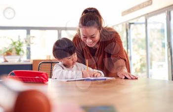 Asian Mother Helping Home Schooling Son Working At Table In Kitchen Writing In Book