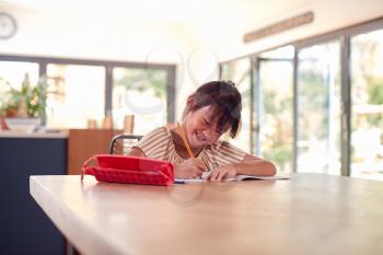 Young Asian Girl Home Schooling Working At Table In Kitchen Writing In Book