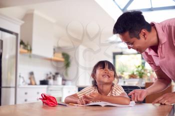 Asian Father Helping Home Schooling Daughter Working At Table In Kitchen Writing In Book