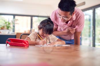 Asian Father Helping Home Schooling Daughter Working At Table In Kitchen Writing In Book