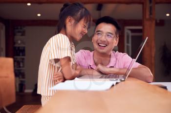 Asian Father Helping Home Schooling Daughter Working At Table In Kitchen On Laptop