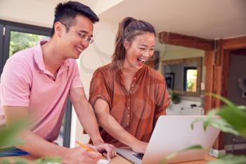 Smiling Couple Sitting At Table At Home Reviewing Domestic Finances Using Laptop