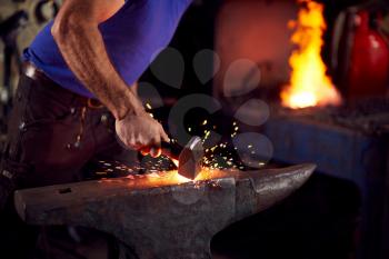 Close Up Of Male Blacksmith Hammering Metalwork On Anvil With Sparks And Blazing Forge In Background