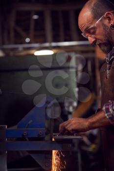 Male Blacksmith Shaping Metalwork On Belt Sander With Sparks