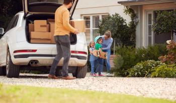 Family Outside New Home On Moving Day Unloading Boxes From Car As Son With Skateboard Helps