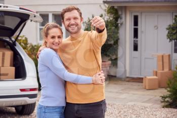 Portrait Of Couple Holding Keys Outside New Home On Moving Day Unloading Boxes From Car