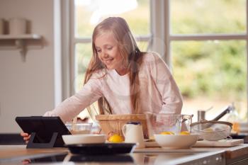 Girl Wearing Pyjamas Making Pancakes In Kitchen At Home Following Recipe On Digital Tablet