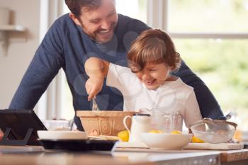 Father And Son In Pyjamas Making Pancakes In Kitchen At Home Following Recipe On Digital Tablet