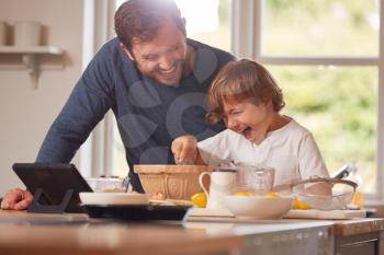 Father And Son In Pyjamas Making Pancakes In Kitchen At Home Following Recipe On Digital Tablet