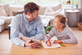 Father Helping Daughter Sitting At Table With Digital Tablet Home Schooling During Health Pandemic
