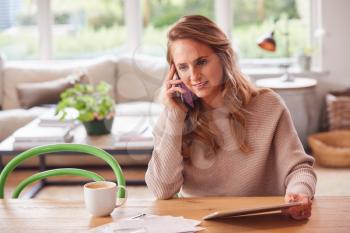 Smiling Woman On Phone Call Sitting At Table At Home With Digital Tablet Reviewing Domestic Finances