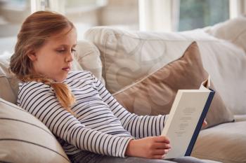 Young Girl Relaxing On Sofa At Home Reading Book