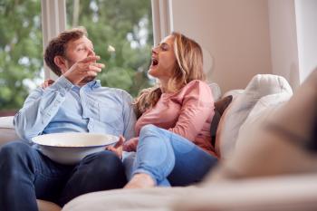 Couple On Date Night Sitting On Sofa At Home Laughing And Catching Popcorn In Mouths