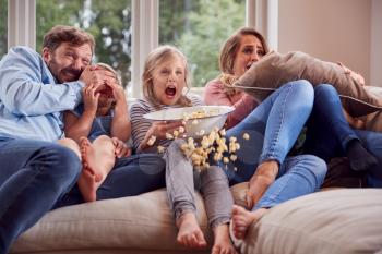 Frightened Family Sitting On Sofa At Home Watching Horror Movie On TV With Popcorn
