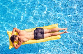 Overhead Shot Of Man In Swim Shorts Floating On Air Bed On Summer Vacation In Outdoor Swimming Pool