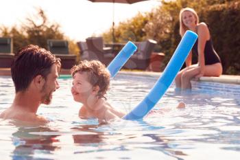 Family In Outdoor Pool On Summer Vacation Teaching Son To Swim With Noodle Swimming Aid
