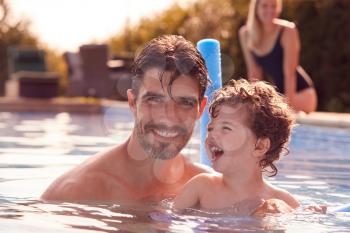 Family In Outdoor Pool On Summer Vacation Teaching Son To Swim With Noodle Swimming Aid