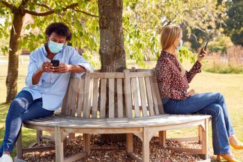 Couple Wearing Masks Meeting In Outdoor Park During Health Pandemic Looking At Mobile Phones