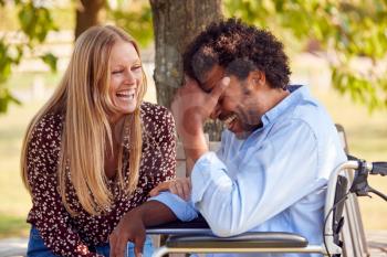Laughing Mature Couple With Man Sitting In Wheelchair Talking In Park Together