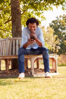 Mature Man Sitting On Bench Under Tree In Summer Park Using Mobile Phone