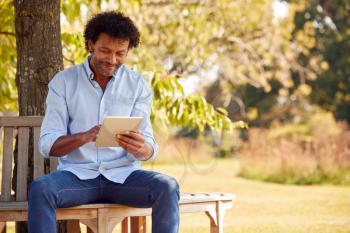 Mature Man Sitting On Bench Under Tree In Summer Park Using Digital Tablet