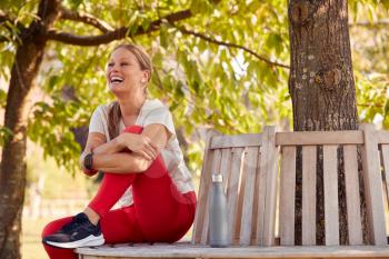 Smiling Woman Wearing Smart Watch And Fitness Clothing Sitting On Seat Under Tree