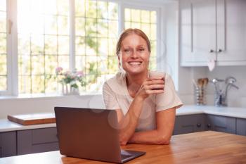 Portrait Of Mature Woman Wearing Fitness Clothing At Home In Kitchen Logging Activity On Laptop