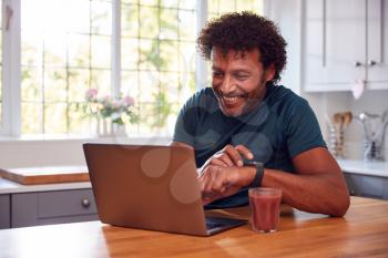 Mature Man Wearing Fitness Clothing At Home Logging Activity From Smart Watch Onto Laptop