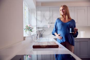 Smiling Pregnant Woman Standing By Kitchen Window Touching Stomach Relaxing With Hot Drink