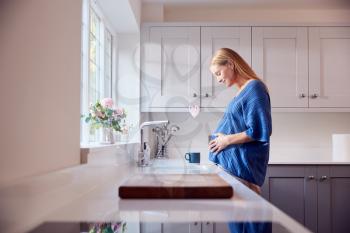 Smiling Pregnant Woman Standing By Kitchen Window Touching Stomach Relaxing With Hot Drink