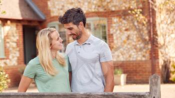 Portrait Of Smiling Couple Standing By Gate Outside Home In Countryside Together