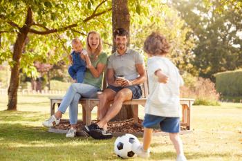 Family With Baby Girl Having Fun In Park Playing Football And Sitting On Seat Under Tree