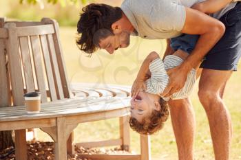 Father With Son Having Fun In Park Holding Him Upside Down Against Leafy Background