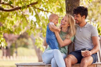 Family With Baby Daughter Sitting On Seat Under Tree In Summer Park Together