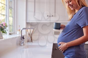 Close Up Of Pregnant Woman Standing By Kitchen Window Touching Stomach Relaxing With Hot Drink