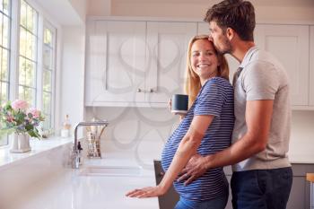 Couple In Kitchen With Man Hugging Pregnant Woman As She Drinks Cup Of Coffee