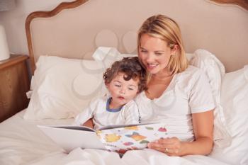 Mother In Bedroom Looking At Picture Book With Young Son Wearing Pyjamas