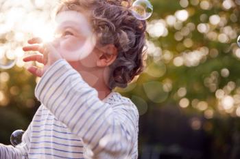 Young Boy Having Fun In Garden Chasing And Bursting Bubbles