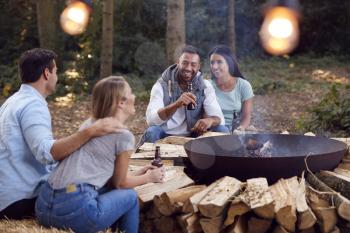 Group Of Friends Camping Sitting By Bonfire In Fire Bowl Drinking Beer Together