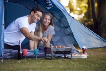 Couple In Tent Camping Sitting By Barbecue Grilling Sausages