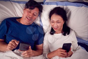 Mature Asian Couple In Wearing Pyjamas Lying In Bed Looking At Mobile Phones Together