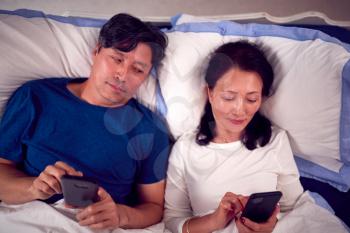 Mature Asian Couple In Wearing Pyjamas Lying In Bed Looking At Mobile Phones Together