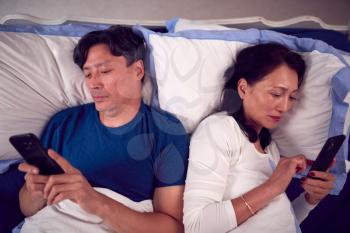 Mature Couple In Wearing Pyjamas Addicted To Using Mobile Phones Lying In Bed And Not Communicating