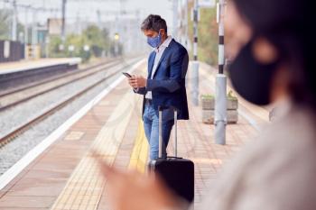 Business Commuters On Railway Platform With Mobile Phones Wearing PPE Face Masks During Pandemic