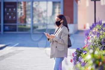 Businesswoman Wearing Mask Walking To Office Looking At Mobile Phone In Health Pandemic