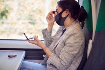 Businesswoman On Train Putting On Makeup Whilst Wearing PPE Face Mask During Health Pandemic