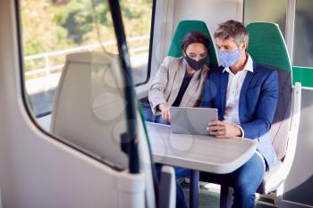 Businesswoman And Businessman On Train Using Digital Tablet Wearing PPE Face Mask During Pandemic