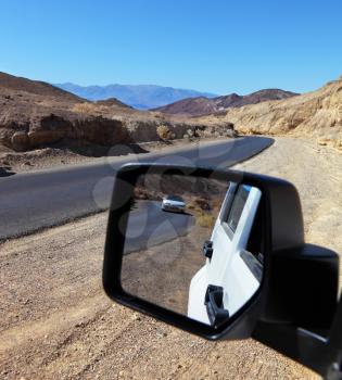 An asphalt road in Death Valley. On the side is a white car. In his review mirror driving affects road and the other white car